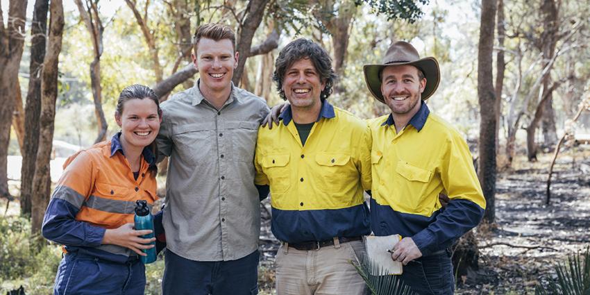 The image shows a group of smiling people in outdoor work attire, standing together in a natural setting.