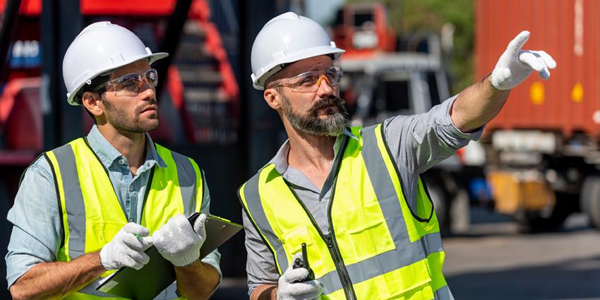 The image shows two workers wearing hard hats, safety vests, and gloves on a construction site, with one holding a clipboard while the other points ahead.