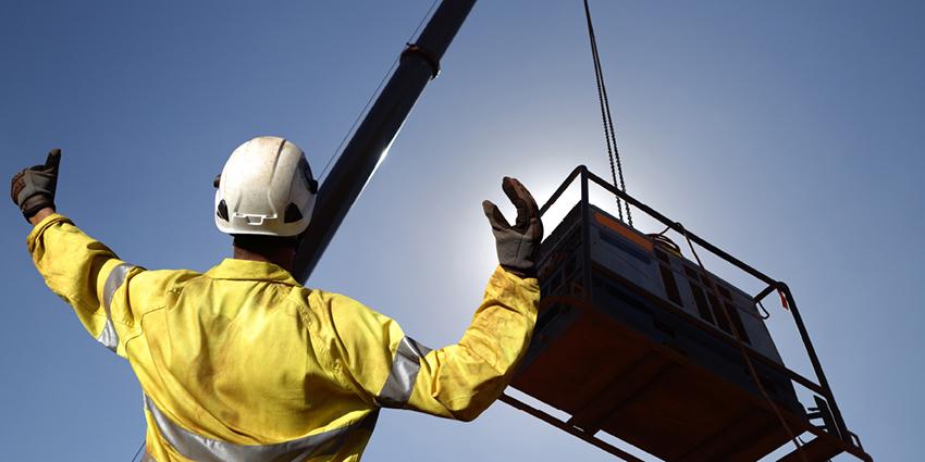 The image shows a worker in high-visibility clothing and a hard hat signaling to a crane operator as a load is lifted, illustrating safe practices in high-risk lifting operations.