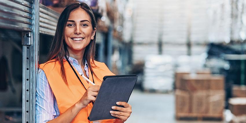 The image shows a smiling worker in a high-visibility vest holding a tablet in a warehouse setting, suggesting efficient management or updating of details and records in a logistics environment.