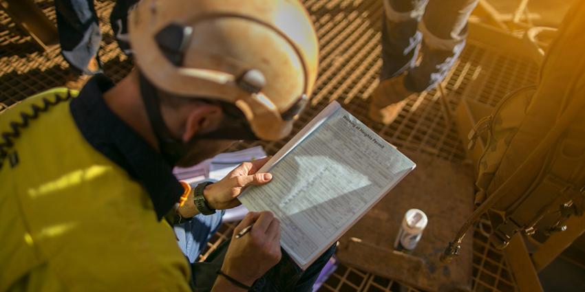 The image shows a worker in safety gear reviewing a checklist or report on a clipboard, likely assessing hazards and documenting safety measures on-site.