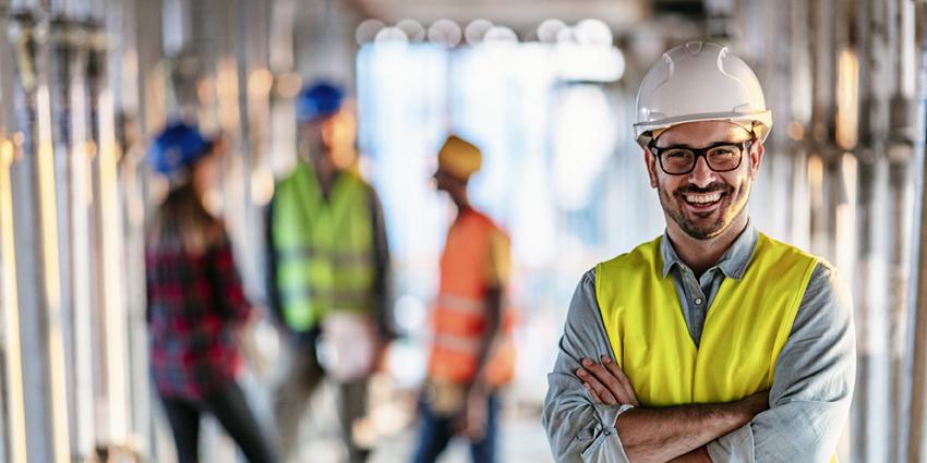 The image shows a construction site setting with a focus on a smiling man wearing a white hard hat, glasses, and a high-visibility safety vest. He has his arms crossed and appears to be confident and approachable. In the background, other workers, also in safety gear, are slightly out of focus, emphasizing the main subject and the workplace safety context. This image conveys a sense of professionalism and commitment to health and safety in a work environment.