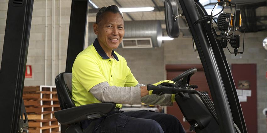 The image shows a smiling worker operating a forklift inside an industrial setting, highlighting a focus on high-risk work requiring specialized skills and safety precautions.