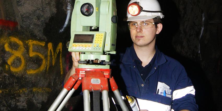 The image shows a mining surveyor wearing a hard hat with a headlamp, operating surveying equipment in an underground setting