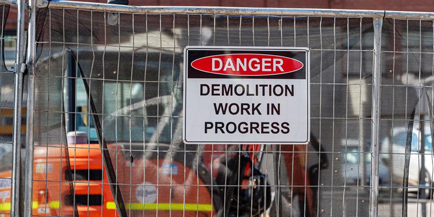 The image shows a warning sign on a fence that reads "DANGER: DEMOLITION WORK IN PROGRESS"