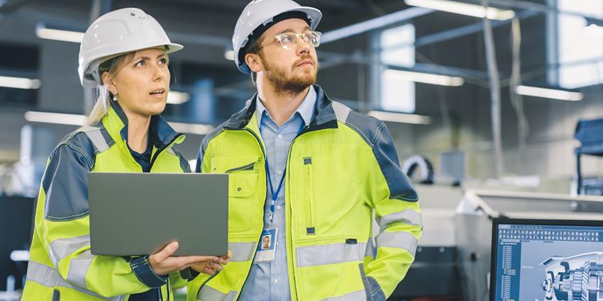 The image shows two professionals in high-visibility jackets and hard hats in an industrial setting, one holding a laptop as they look attentively at something off-frame.