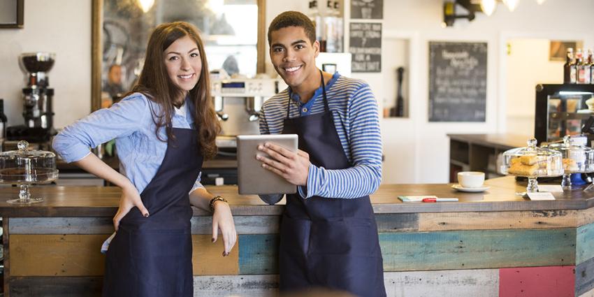The image shows two smiling café employees wearing aprons, standing behind a counter and holding a tablet