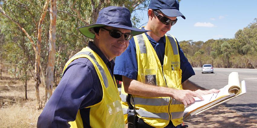 The image shows two WorkSafe inspectors in high-visibility vests, reviewing a document outdoors