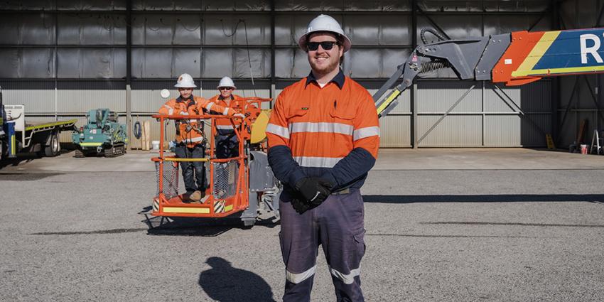 The image shows a worker in high-visibility clothing and a hard hat standing in front of industrial equipment, with two other workers in a lifting platform behind him