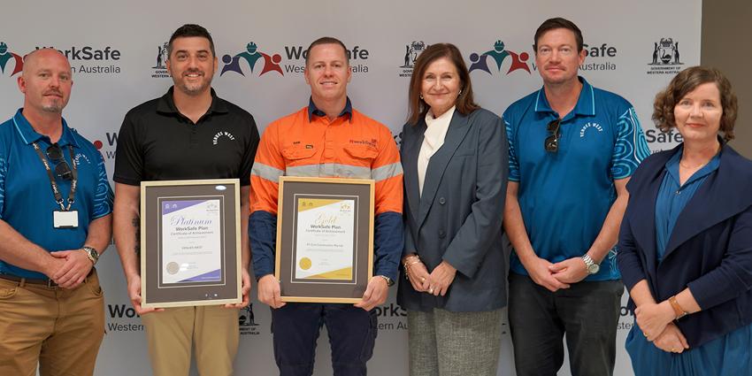 The image shows a group of people standing in front of a WorkSafe Western Australia banner, holding a Platinum and Gold WorkSafe Plan Certificate of Achievement